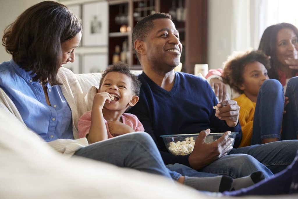 The image of a three-generation family sitting on the sofa in a living room, watching TV and eating popcorn.