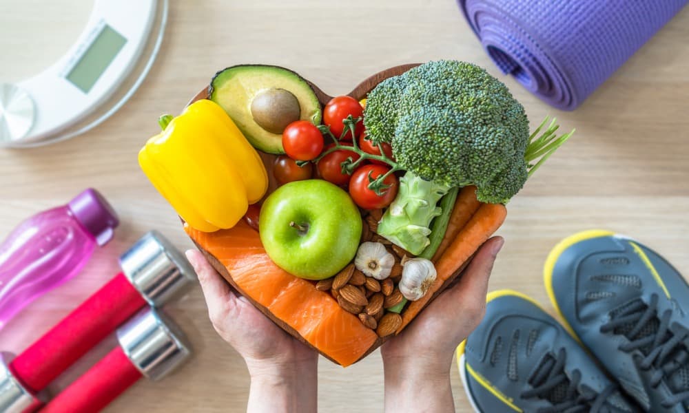 A heart-shaped plate of healthy vegetables, fruits, and nuts surrounded by exercise equipment such as a yoga mat, running shoes, and dumbbells.